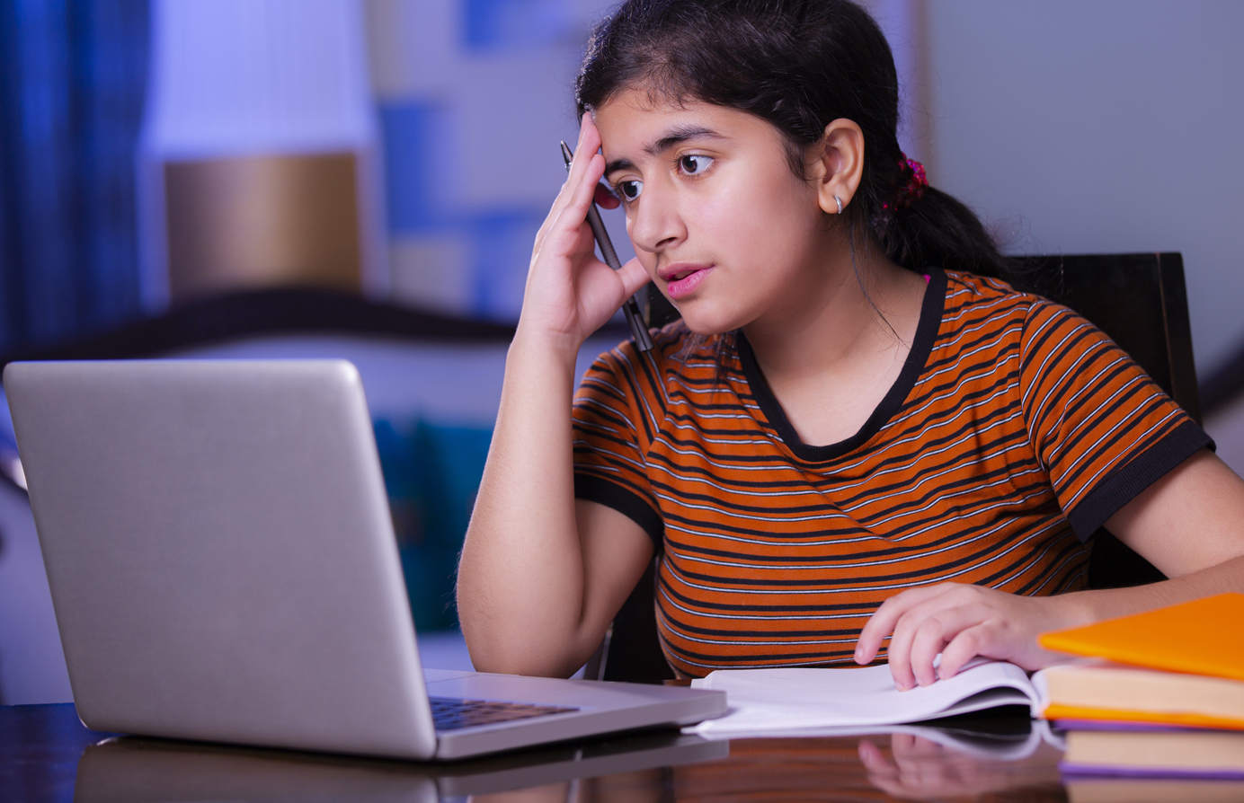 A young girl in a striped shirt sits at a desk looking stressed, with one hand on her head, talking on the phone while staring at her laptop and an open notebook, studying or doing homework.